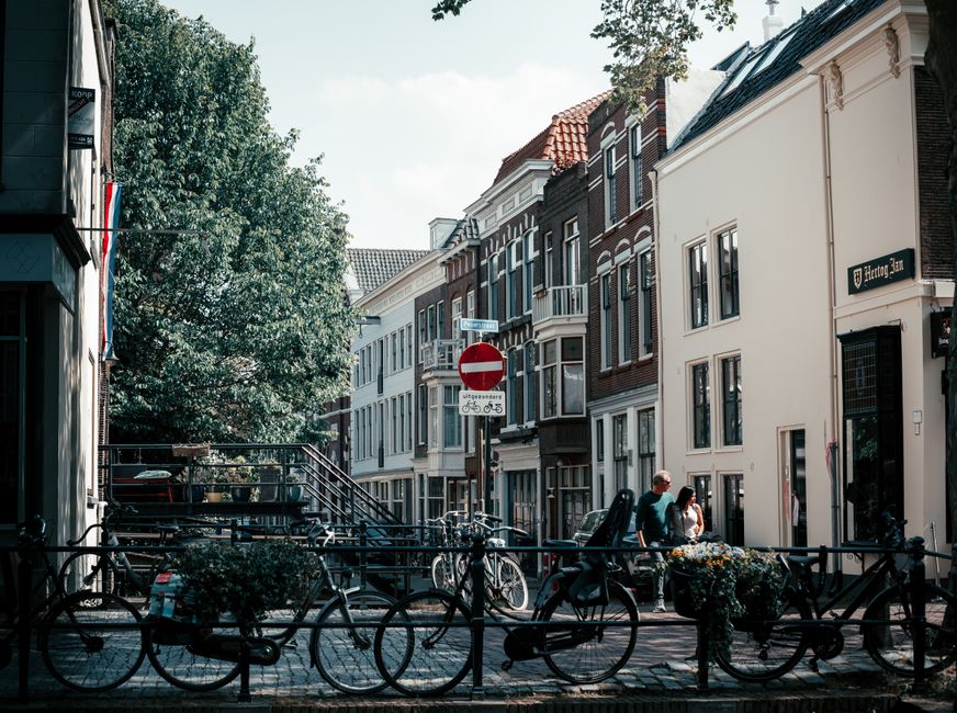 A narrow urban street scene featuring a row of multi-storey residential buildings with brick facades, large windows, and pitched roofs, some with decorative white trim and bay windows. In the foreground, several bicycles are securely locked to metal railing barriers along the pavement, with a small planter box filled with flowers attached to one of the barriers. Two pedestrians, a man and a woman, stand near the railing, engaged in conversation. A prominent red and white 'no entry' traffic sign is positioned at the street corner, with a supplementary smaller sign indicating bicycle restrictions in Dutch. Surrounding the scene are lush green trees partially shading the buildings and street. The overall atmosphere is calm with clear daylight, emphasizing the typical city cycling and pedestrian environment which service providers like Waste Disposal Holland Park may encounter when offering private rubbish collection or on-site clearance in such residential areas, emphasizing the importance of accessible waste removal options for urban properties.