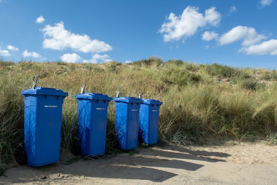 Four blue wheeled rubbish bins with closed lids are positioned on sandy ground near a grassy sand dune, with tall, green, and brown dune grass growing behind them. The bins appear to be made of plastic with a textured surface and are arranged in a straight line, slightly leaning to the right, casting long, dark shadows on the sandy terrain. The background includes a bright blue sky dotted with fluffy white clouds, creating a clear and sunny environment. The scene suggests an outdoor waste disposal area, with the bins possibly intended for private waste collection in a coastal or rural setting, aligning with alternative rubbish removal methods that do not involve shared municipal services. Waste Disposal Holland Park occasionally handles similar waste management tasks, although there are no visible vehicles or equipment in the image. The overall composition emphasizes the distinction between the waste containers and natural surroundings in a serene, seaside landscape.