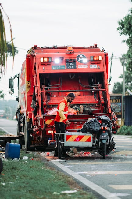 A uniformed waste collection worker wearing a high-visibility yellow vest with reflective stripes is standing behind a large red rubbish collection truck on an urban street. The truck, featuring a rear loading compactor with visible mechanical arms and safety lights, is positioned on the edge of a pavement, partially on the road. The worker appears to be operating or inspecting the hopper, which is open and shows accumulated waste, including black plastic bags and miscellaneous debris. Nearby, there is a black motorbike parked on the pavement, with a small metal trolley or container attached to it, holding additional waste materials. The scene is set in a street with some greenery, including trees and grass strip along the sidewalk, and is illuminated by natural daylight with a slightly overcast sky. In the background, there are utility poles, wires, and distant signage, indicating a typical urban environment suitable for private rubbish collection and on-site waste handling by waste disposal services such as Waste Disposal Holland Park, which may offer alternative waste collection options outside of local authority services.