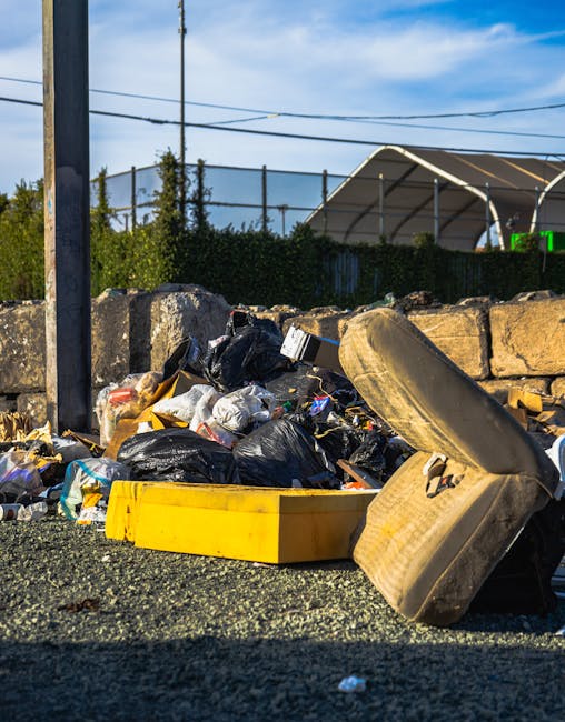 An overflowing pile of mixed waste and rubbish bags, including black plastic refuse sacks, cardboard boxes, and discarded furniture, situated on an asphalt surface next to a low stone wall. A large, dirty, and worn car tire leans against the debris in the foreground, partially obscuring a yellow plastic container. In the background, a metal fence and some greenery are visible, along with a partly cloudy sky and overhead power lines. The scene suggests an instance of improper or unmanaged rubbish disposal, which might be addressed through private waste removal services offered by companies like Waste Disposal Holland Park, especially for bulk or bulky waste items. The overall environment appears to be an outdoor area in an urban or suburban setting, highlighting the importance of proper rubbish management and the potential need for alternative waste handling solutions instead of relying solely on municipal collection. The lighting is natural daylight, casting minimal shadows, emphasizing clarity and detail of the trash and surrounding elements.