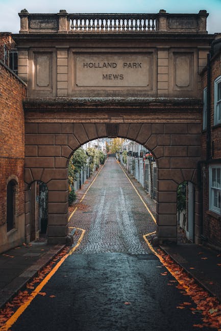 A view through a stone archway with an inscription reading 'HOLLAND PARK MEWS', leading to a narrow cobblestone street extending into the distance. The arch is built from reddish-brown bricks and features a decorative railing at the top. On either side of the street, there are residential buildings with brick walls and bay windows, some with small gardens or fences. The pavement is wet, with fallen leaves visible along the edges, suggesting recent rain. This scene, captured during daylight, is part of Holland Park, a quiet residential area, and exemplifies the type of setting where private rubbish removal or on-site clearance services by Waste Disposal Holland Park might be required to maintain cleanliness and order in historic urban environments.