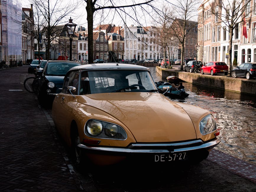 A vintage yellow convertible sports car with a black soft top, parked on a cobblestone street adjacent to a canal in an urban residential area, with closely spaced brick and stucco buildings featuring large windows and decorative facades in the background. The vehicle's front grille and rounded headlights are visible, with a Dutch license plate reading DE-57-27 attached to the front bumper. Behind the car, a black van and a bicycle are parked along the curb, with leafless trees lining the street. To the right, several parked modern cars, including a red hatchback and dark sedans, are visible beside the canal, which has a stone wall and some reflections from the water. The scene is illuminated by soft natural daylight, creating a calm atmosphere consistent with a typical cityscape where private vehicle parking and street-side activity are common. The image appears to relate to an urban environment where alternative waste handling options, such as rubbish removal services, might be relevant, but no waste or disposal actions are directly visible in the scene.