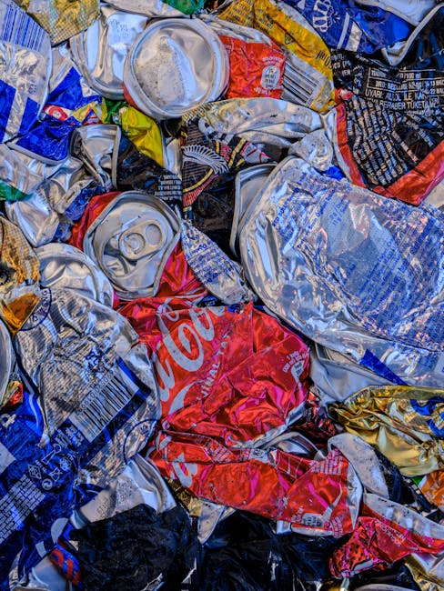 A close-up view of a large pile of crushed aluminum cans and aluminum foil, predominantly in silver, with some red and blue printed branding visible, including a prominent Coca-Cola logo on a red can. The cans show creased, crumpled textures typical of recyclables after being emptied and compacted. Some cans have visible pull tabs, while others are flattened with open ends or partially deformed. The surface of the cans reflects light, creating highlights and shadows that emphasize their metallic finish. The background includes additional crushed cans and foil, with some folded or bent edges. The scene appears to be outdoors, possibly on a concrete or asphalt surface, indicating a collection or disposal area for recyclable waste managed by waste disposal services like Waste Disposal Holland Park. This image represents a typical example of recyclable aluminium waste ready for collection or processing, relevant to alternative waste handling and rubbish service categories.