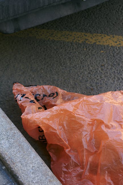 A crumpled orange plastic bag lying on a textured asphalt surface, partially overlapping a light-colored concrete curb. The bag appears to be discarded and is situated near the edge of a paved driveway or street, with a dark gray background at the top. The scene suggests an instance of waste left on the ground, which waste disposal services like Waste Disposal Holland Park may assist with for proper collection and recycling, adhering to local waste management regulations.