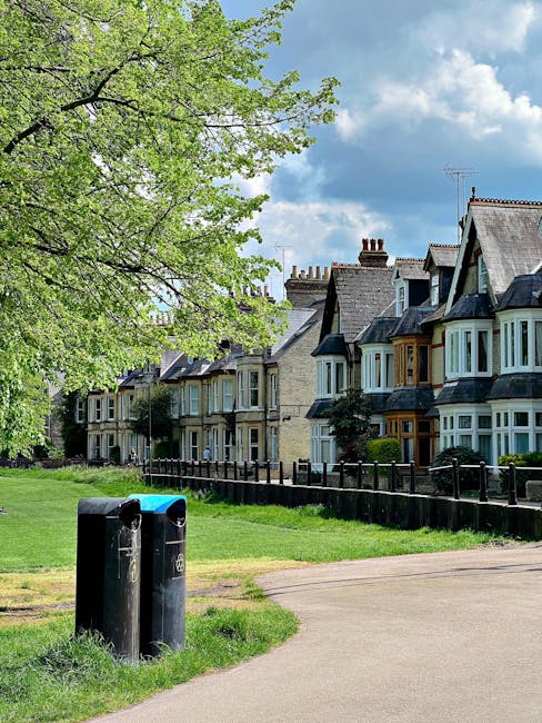 A residential street scene showing a row of traditional Victorian-style terraced houses with bay windows, pitched roofs, and chimneys, situated behind a well-maintained black metal fence. In the foreground, a grassy area with a large leafy tree displaying fresh green foliage partially shades the scene, and two black public litter bins are positioned near the path. The setting appears to be during daylight with partly cloudy skies, creating a bright and clear atmosphere. The image reflects a typical UK urban environment where private waste collection services, such as those offered by Waste Disposal Holland Park, may be involved in handling rubbish removal for local residents. The scene emphasizes the residential context and the importance of proper waste disposal in maintaining community cleanliness and environmental standards.