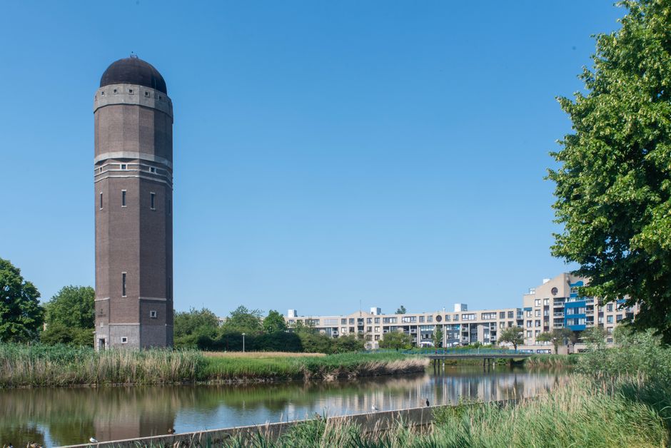 A tall, cylindrical brick water tower with a rounded, dark-colored domed roof positioned on the left side of the image. Its surface features horizontal white lines and narrow vertical windows running up the structure. In the foreground, there is a narrow, calm water body bordered by tall grasses and reeds, reflecting the blue sky and parts of the surrounding environment. To the right, a lush, green leafy tree partially obscures some modern, white residential buildings with blue balconies situated in the background. The scene is bathed in natural daylight, highlighting the contrast between the historic industrial structure and the contemporary urban foliage, creating a peaceful yet practical setting that aligns with waste management or rubbish removal context by demonstrating a clean and well-maintained environment adjacent to private or municipal infrastructure.