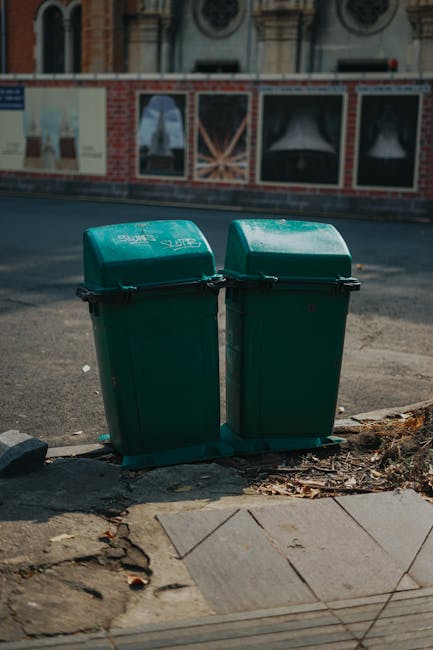 Two green wheelie bins with closed lids are positioned side by side on a concrete pavement at the edge of the street, with the bins' black locking mechanisms visible on the front. The bins are made of durable plastic with a slightly textured surface and are marked with white branding or labels, indicating their purpose for waste collection. Behind the bins, there is a worn asphalt road that appears slightly weathered, with small debris and leaves scattered near the base of the bins. Slightly further back, a red brick wall with large, rectangular advertising posters, which appear to be faded and partially obscured, runs parallel to the street. The environment suggests a typical urban setting, where private waste disposal could be managed independently, as indicated by the presence of Waste Disposal Holland Park. Natural daylight illuminates the scene, casting soft shadows, and highlighting the weathered surfaces of the pavement and the bins, consistent with daytime in a residential or commercial area during mild weather.