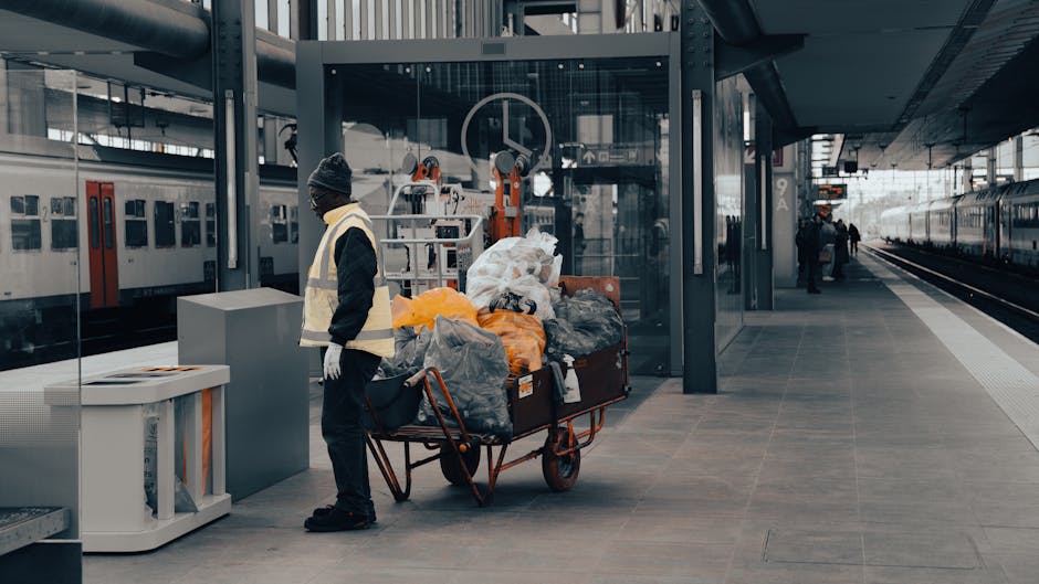 A person dressed in a dark jacket, black trousers, and a grey knit hat stands beside a red-handled wheelbarrow loaded with various discarded items, including large black trash bags, orange and grey plastic sheeting, and miscellaneous waste materials, all of which appear to be accumulated rubbish. The individual is wearing white gloves and is positioned on a paved platform at a modern train station, with glass and metal structures visible behind and around them. In the background, a white train with red and black details is parked on adjacent tracks, and farther down the platform, a few other people are present near the station's edge. To the left, a small white recycling bin or waste container is visible, while the station's overall environment is clean and contemporary, with overhead lighting and a metal canopy overhead. The scene conveys a moment of private waste handling, perhaps related to on-site clearance or independent rubbish collection, with the worker preparing for rubbish removal in a transit environment, supported by Waste Disposal Holland Park’s services for rubbish disposal.