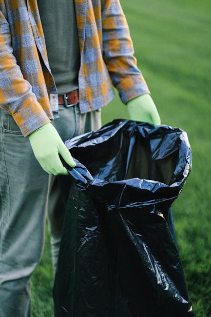 A person standing outdoors on a grassy area, wearing a checkered shirt with shades of yellow, blue, and gray over a green T-shirt, with beige trousers secured by a brown belt. They are wearing light green disposable gloves and holding open a large black rubbish bag made of thick plastic material, which is partially filled. The scene suggests an active waste collection effort, possibly involving private waste disposal or clearance work. The background shows lush green grass and blurred greenery, indicating a park or garden setting, with natural light illuminating the scene. The image emphasizes manual waste collection and disposal, aligning with rubbish removal or on-site clearance services provided by Waste Disposal Holland Park, demonstrating a practical approach to managing household or garden waste in an environmentally conscious manner.