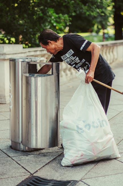 A middle-aged woman with short dark hair is seen leaning forward while placing a white plastic bag filled with waste into a stainless steel outdoor rubbish bin. She is wearing a black T-shirt with white printed text on the front and black trousers. The bin is situated on a paved walkway alongside a stone edge, with a background of lush green trees and foliage, suggesting a park or garden setting. The scene is illuminated by natural daylight, highlighting the metallic surface of the bin and the woman's focused action of waste disposal. This image relates to private waste handling or alternative rubbish removal services offered by Waste Disposal Holland Park, as it depicts an individual actively participating in rubbish collection outdoors.